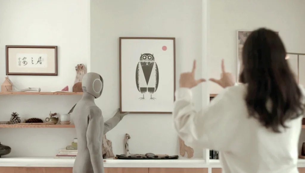 A humanoid robot hanging an owl illustration on a wall as a person gestures towards it.