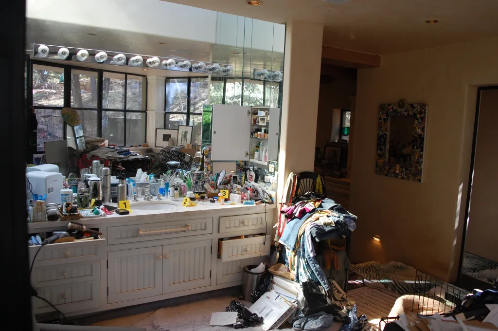 A messy bathroom with a white vanity covered in toiletries, and a pile of clothes next to it.