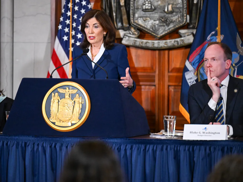 New York Governor Kathy Hochul speaks at a podium with Blake Washington, Budget Director of New York State, listening nearby.