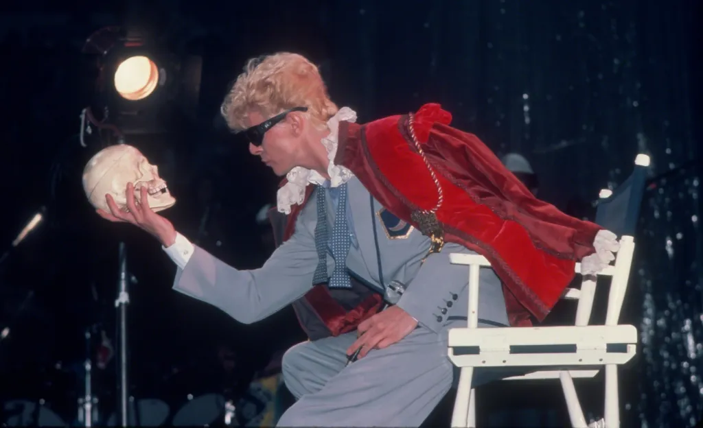 David Bowie performing during the Serious Moonlight Tour, dressed in a grey suit with a red cape and ruffled collar, holding a skull.