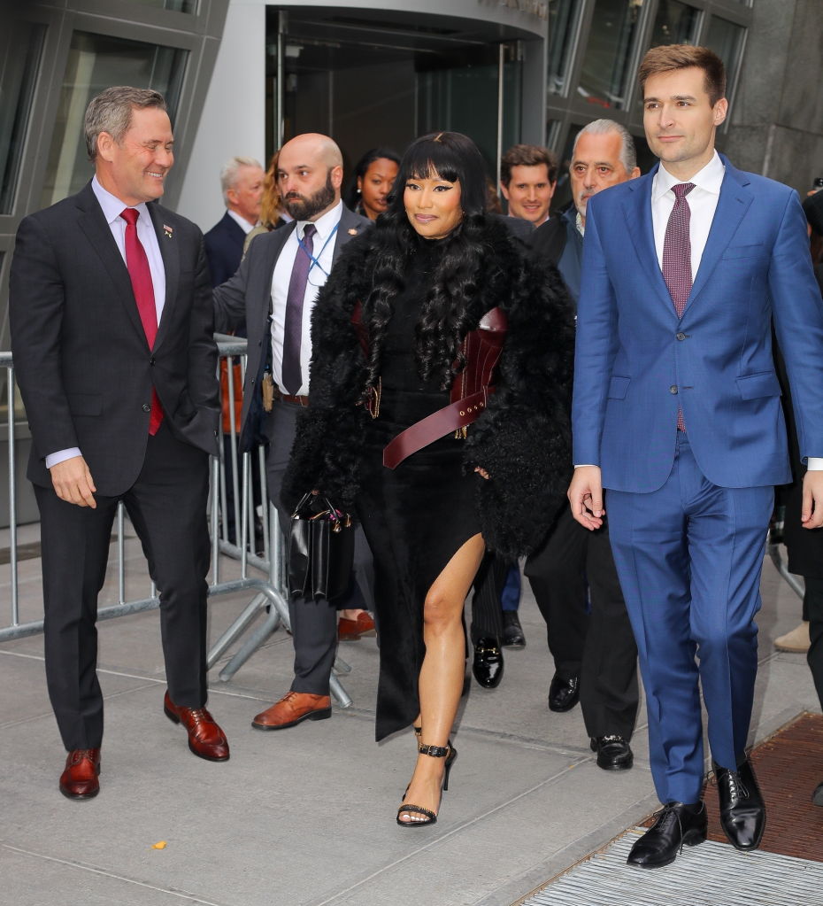 Nicki Minaj arriving at the United Nations in New York, wearing a black dress with a high slit, a fur coat, and strappy heels, accompanied by men in suits.