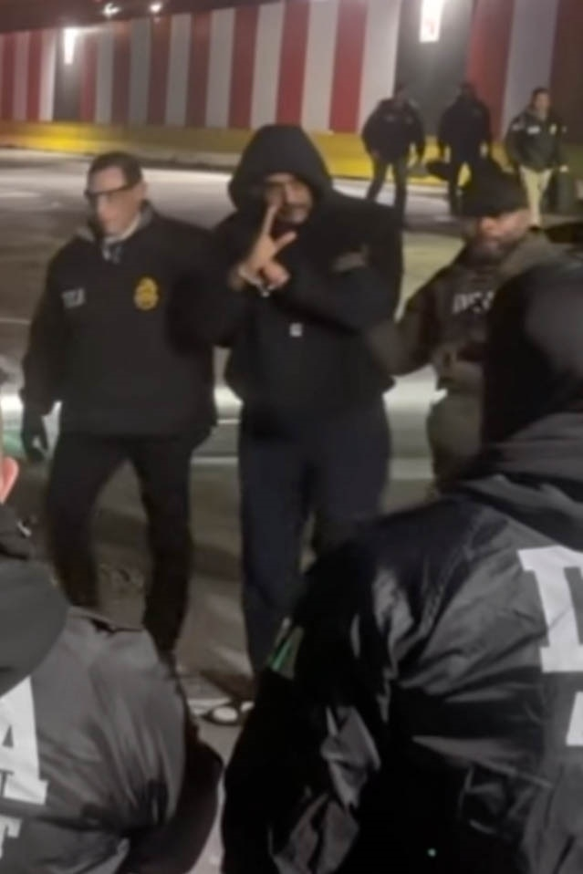 Nicolas Maduro in a dark hoodie and pants, making a hand gesture while surrounded by law enforcement officers.