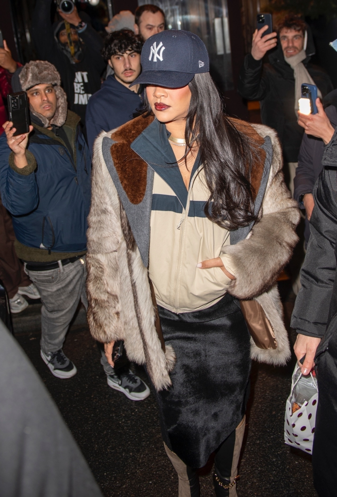 Rihanna exiting her hotel wearing a baseball cap and trench coat, surrounded by a crowd of fans and photographers.