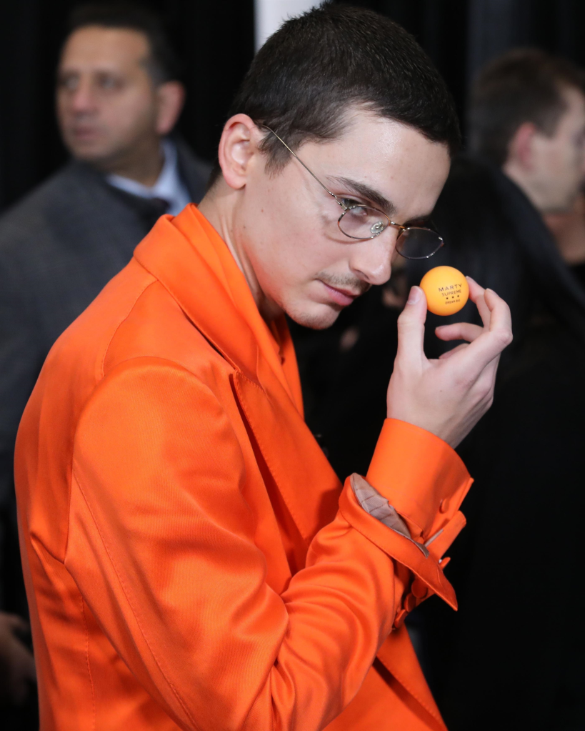 Timothée Chalamet wearing an orange suit, holding an orange ping pong ball with 