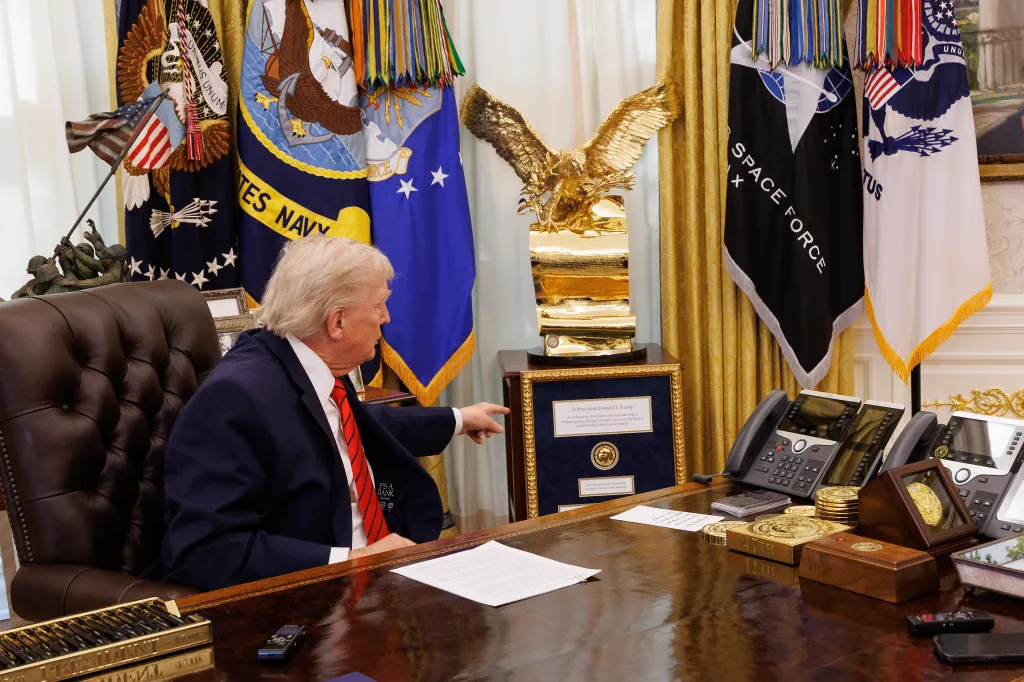 Donald Trump sitting at a desk in the Oval Office, pointing to a framed Nobel Peace Prize.