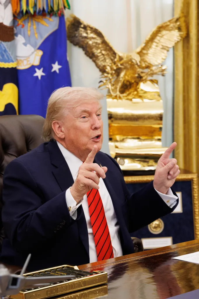 Donald Trump sitting at a desk with an eagle statue and flags behind him, gesturing while speaking during an interview.