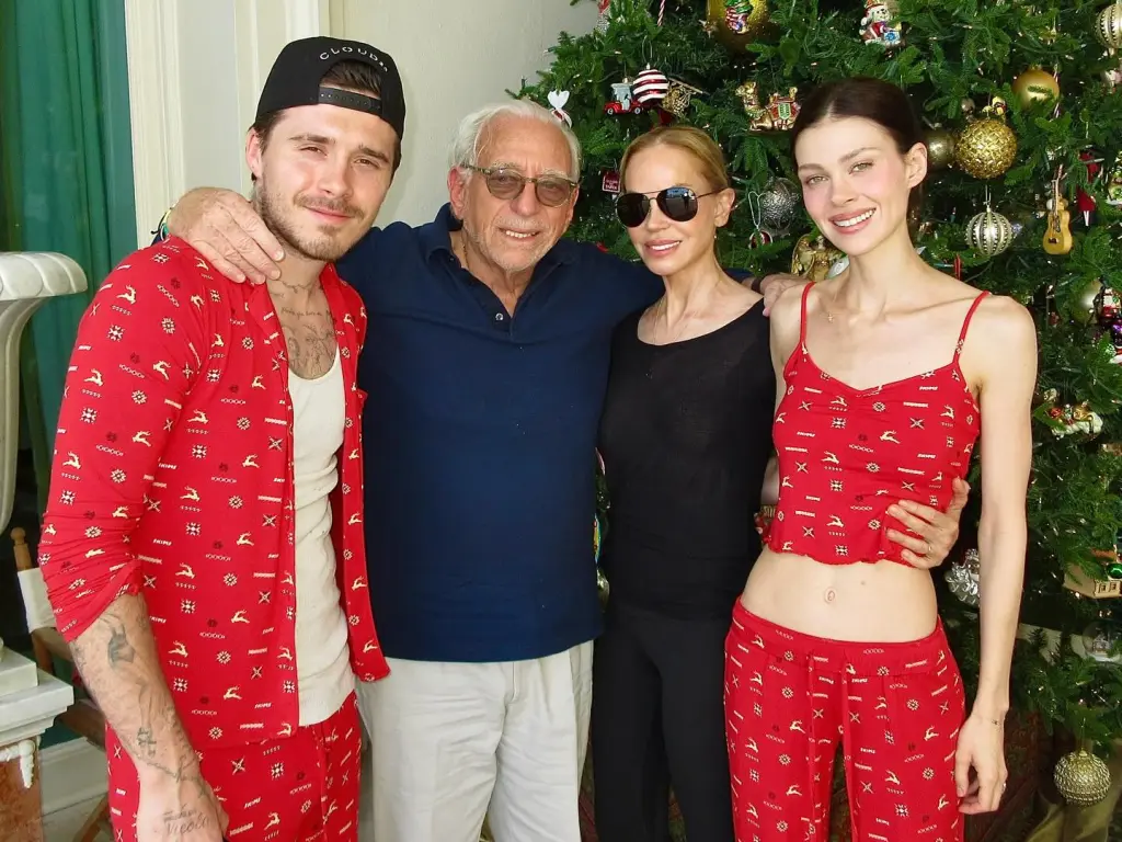 Brooklyn Beckham and Nicola Peltz, in matching red pajamas, with Nelson Peltz, in a blue sweater and cream pants, and Claudia Peltz in a black top and black pants.