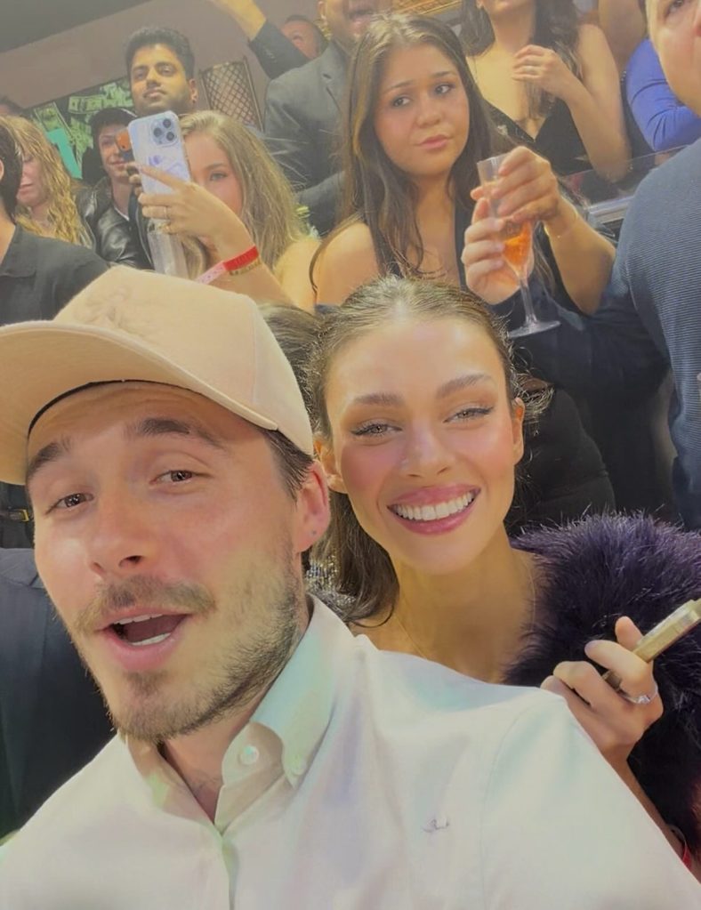 Nicola Peltz and her husband Brooklyn posing for a selfie in a crowd. Brooklyn wears a white shirt and cream baseball cap. Nicola in a purple fluffy jacket.
