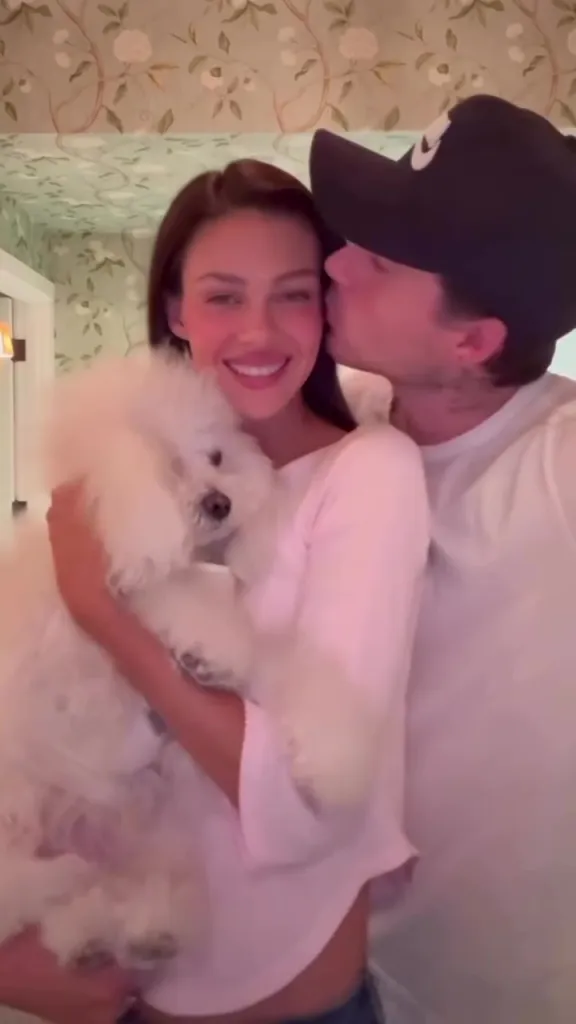 Nicola Peltz holding a white dog while Brooklyn Beckham kisses her cheek.