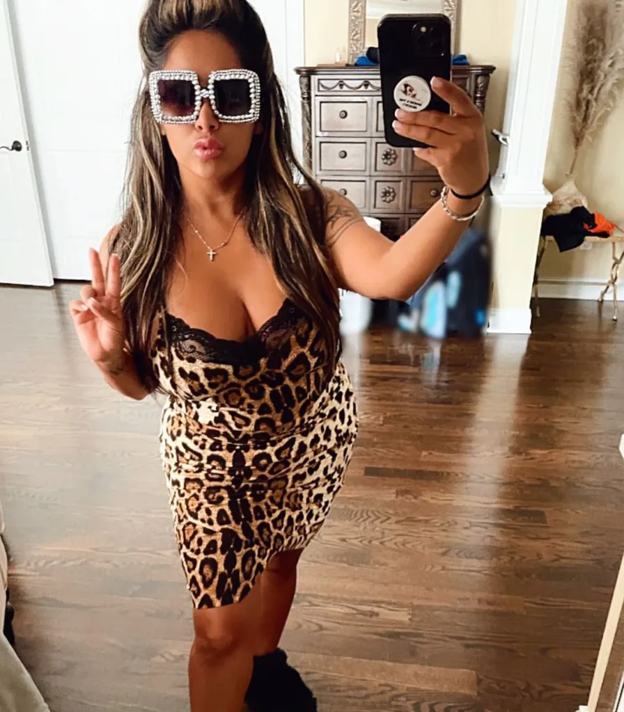 Nicole Polizzi taking a mirror selfie, wearing a leopard print dress and large, rhinestone-studded sunglasses, with her hair in a half-up style and giving a peace sign.