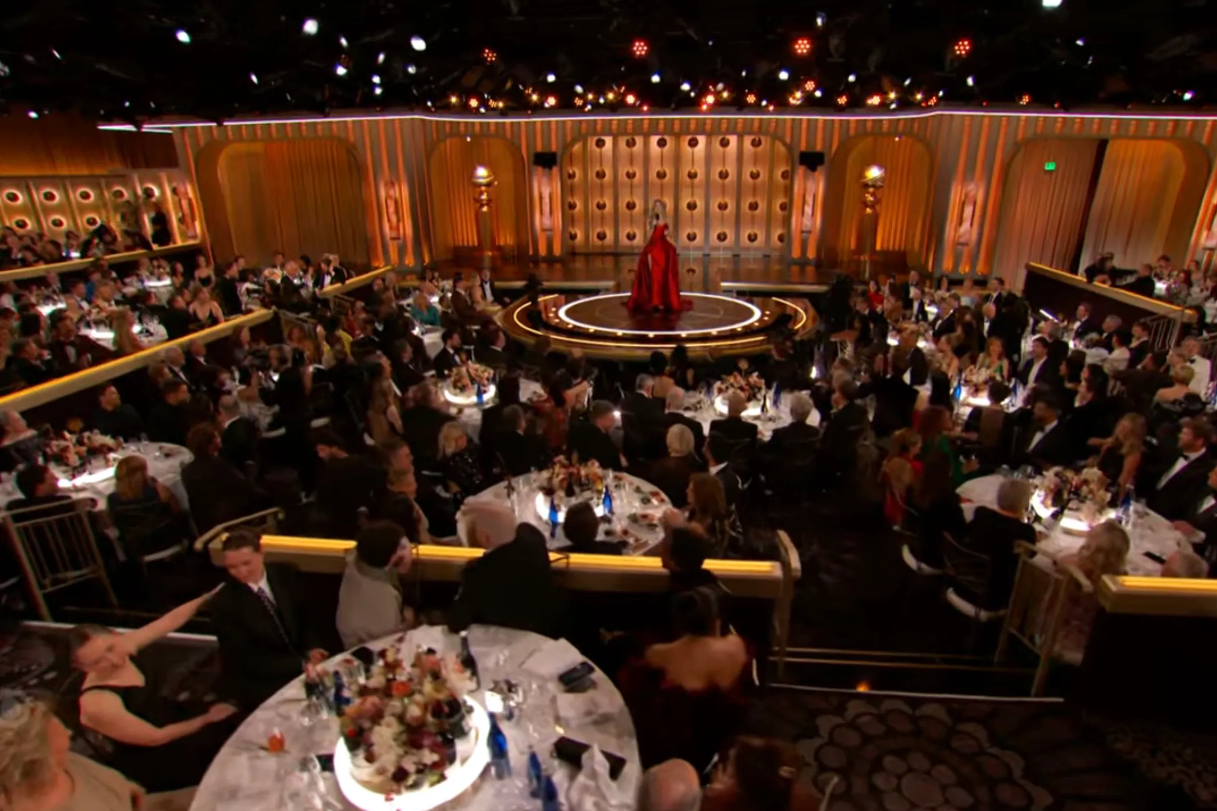 Nikki Glaser in a red dress on stage, addressing guests seated at tables during the 83rd Annual Golden Globe Awards.