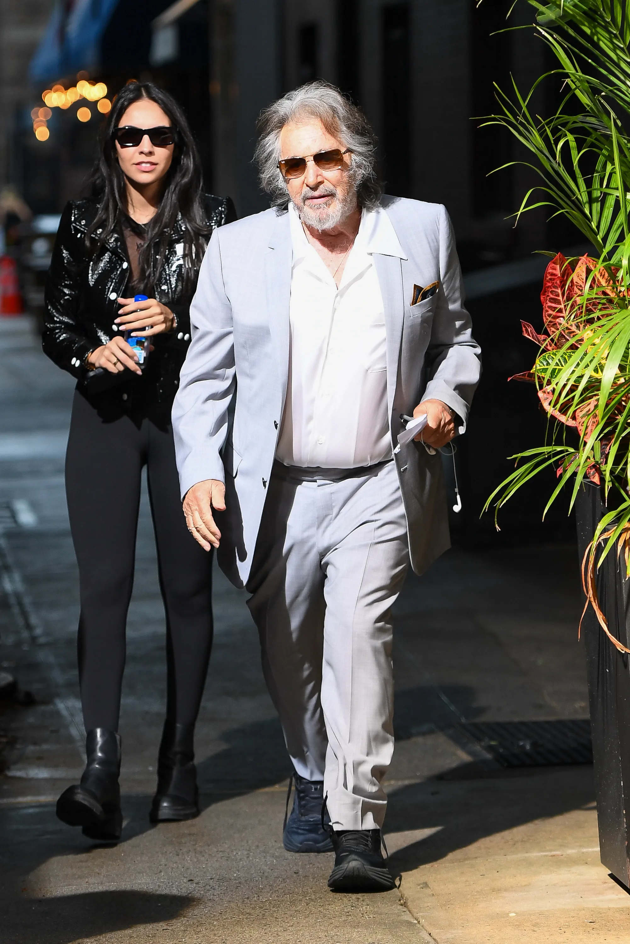 Noor Alfallah walks behind Al Pacino, who wears a light gray suit and sunglasses.