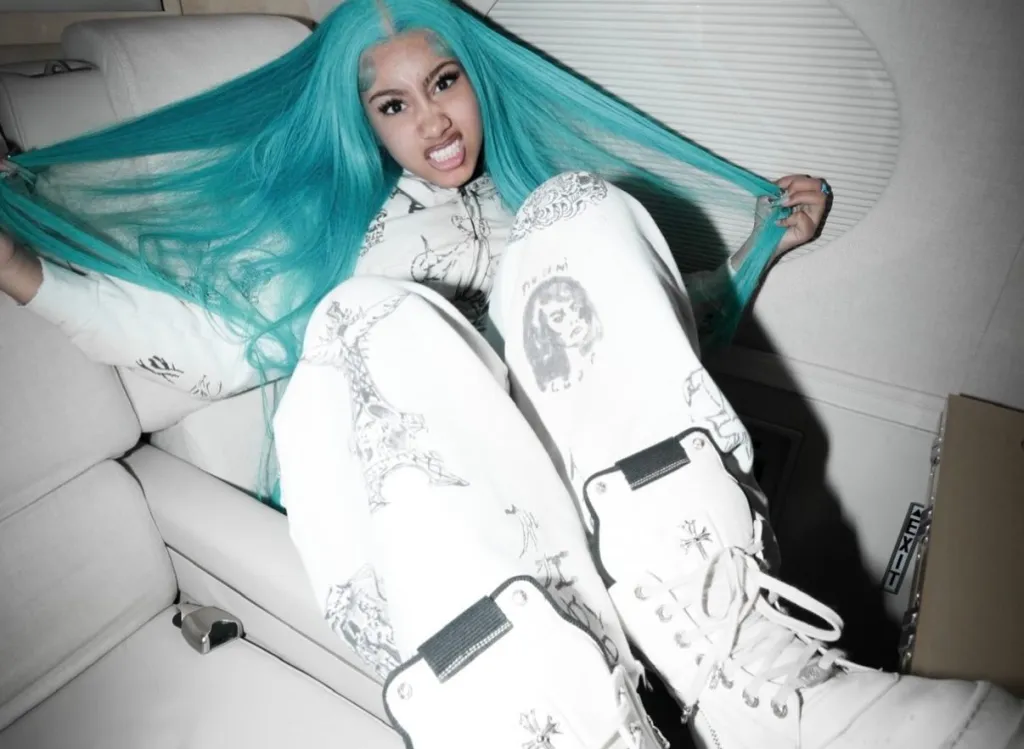 Person with long teal hair and diamond grills wearing white graphic sweatpants and high-top sneakers, sitting on a white couch.
