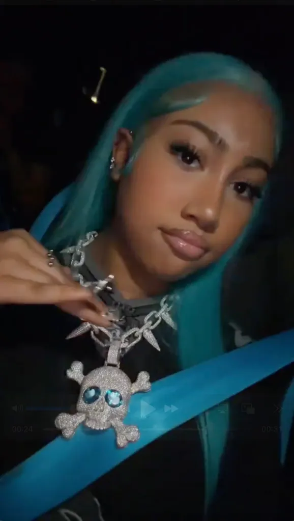 A woman with blue hair, wearing a skull and crossbones necklace, looks into the camera.