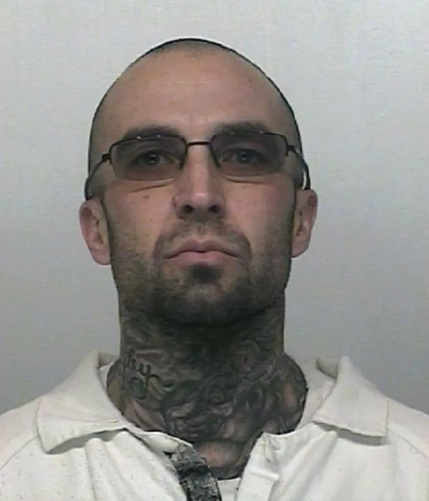 Mugshot of Kody Olsen, a bald man with a beard and neck tattoos, wearing glasses and a white collared shirt.