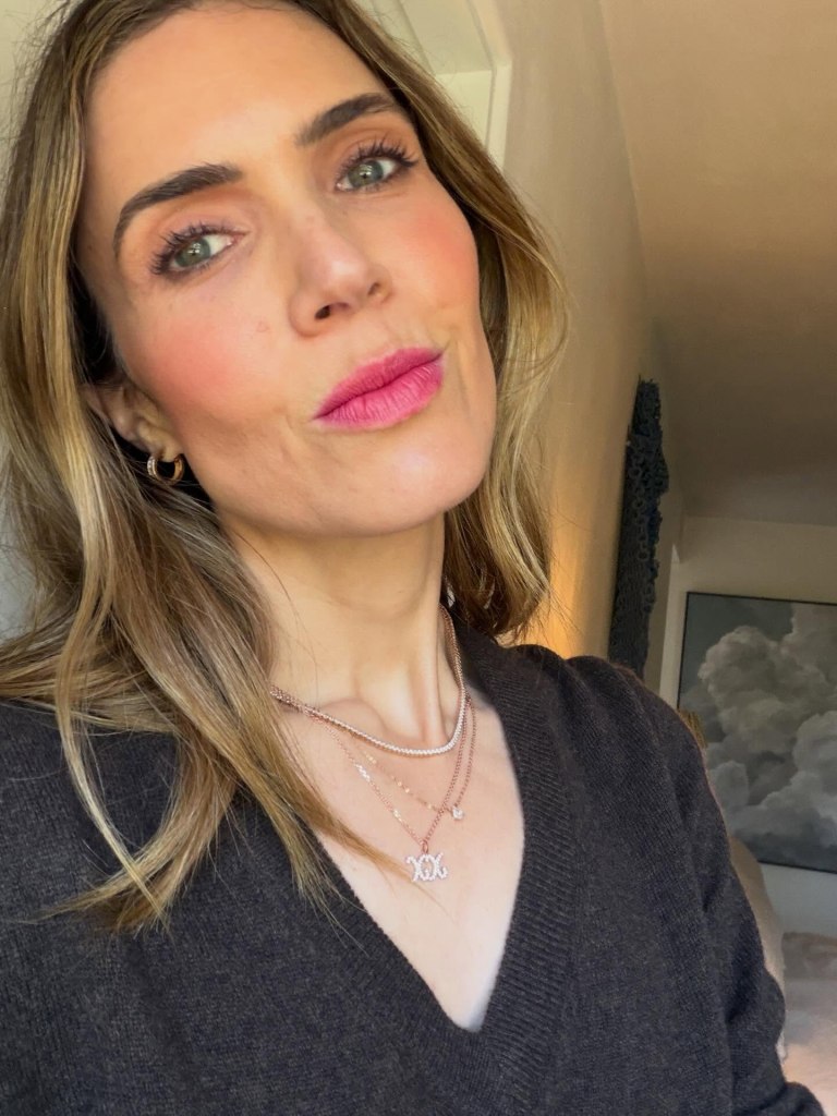 Mandy Moore in a dark V-neck sweater and layered rose gold necklaces, with bright pink blush and lipstick.