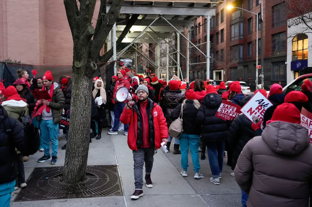 Nurses, wearing red hats and winter clothes, demonstrate outside Mount Sinai West Hospital during a strike, with one nurse speaking into a megaphone.