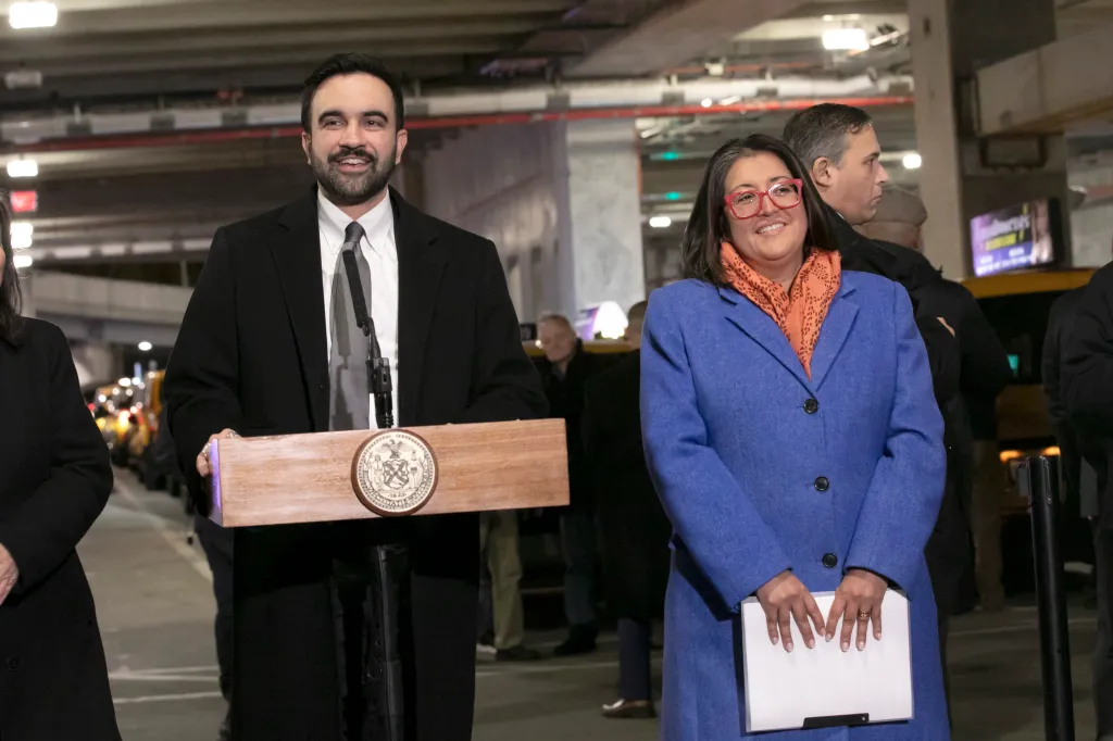 NYC Mayor Zohran Mamdani stands at a podium next to Midori Valdvia at LaGuardia Airport.