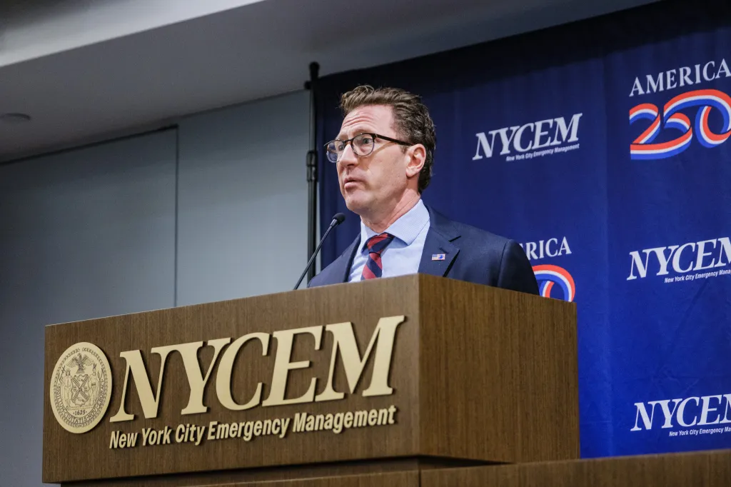 NYCEM Commissioner Zach Iscol speaking at a podium in front of a blue backdrop with the NYCEM logo.