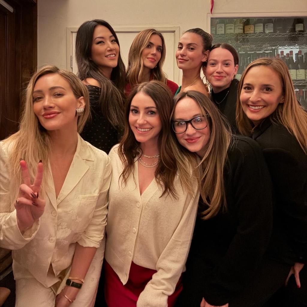 A group of eight women posing together in a dimly lit room with bottles on shelves in the background.