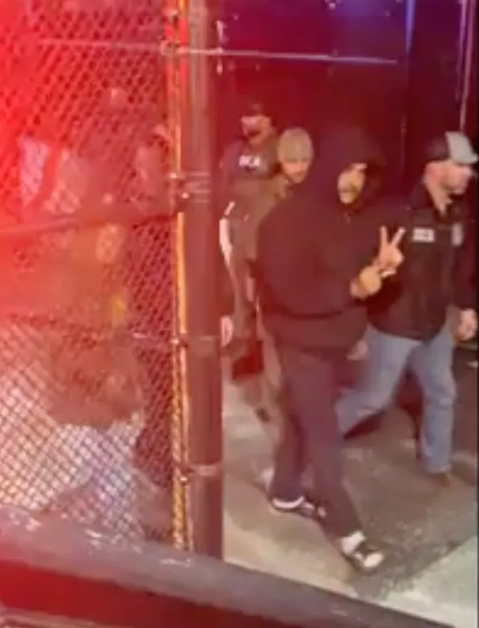 Nicolás Maduro, wearing a black hoodie and slides, gives a peace sign while being escorted by DEA agents.