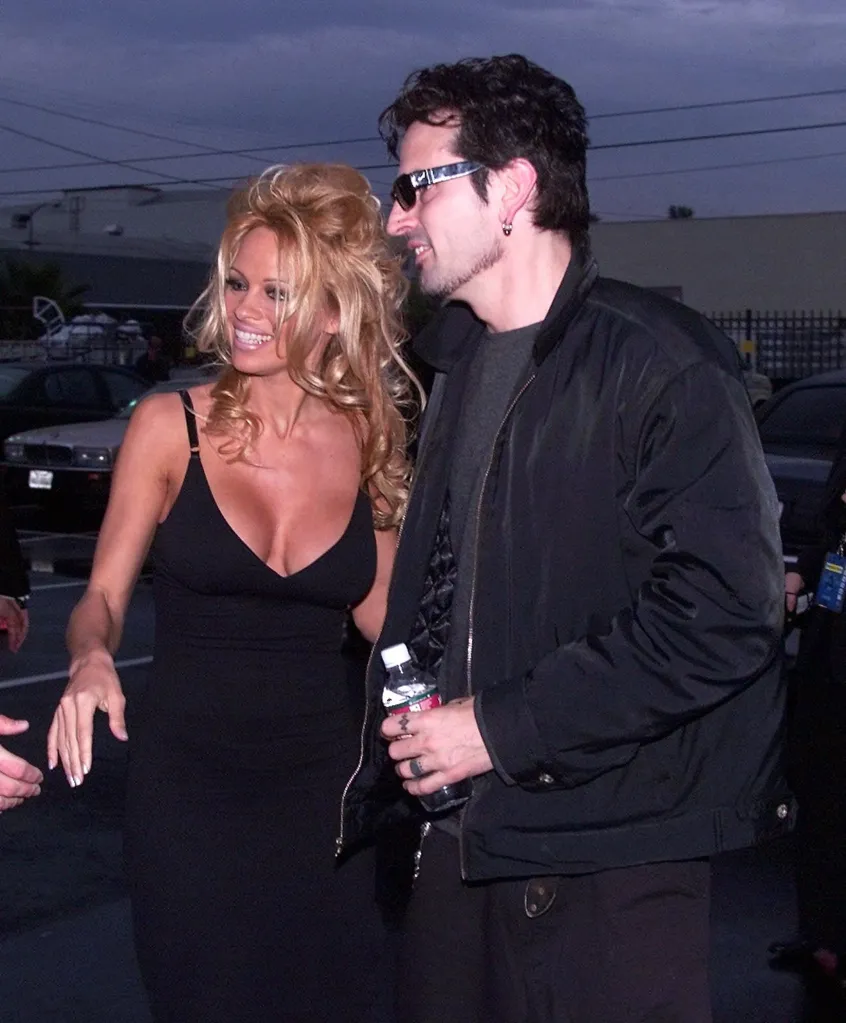Pamela Anderson Lee in a black dress and Tommy Lee in a black jacket and sunglasses smile while walking.