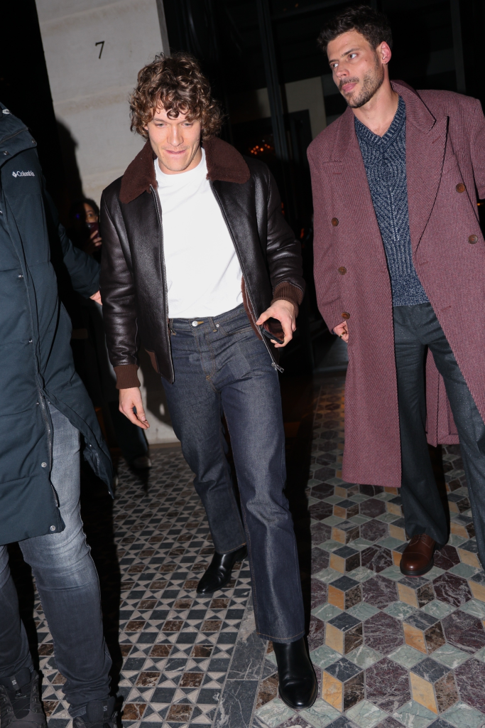 François Arnaud in a black leather jacket and Connor Storrie in a burgundy coat walk outside at night.
