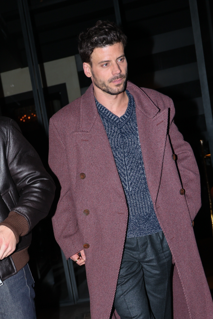 François Arnaud in a burgundy coat, blue sweater, and tailored pants, with Connor Storrie's arm visible.
