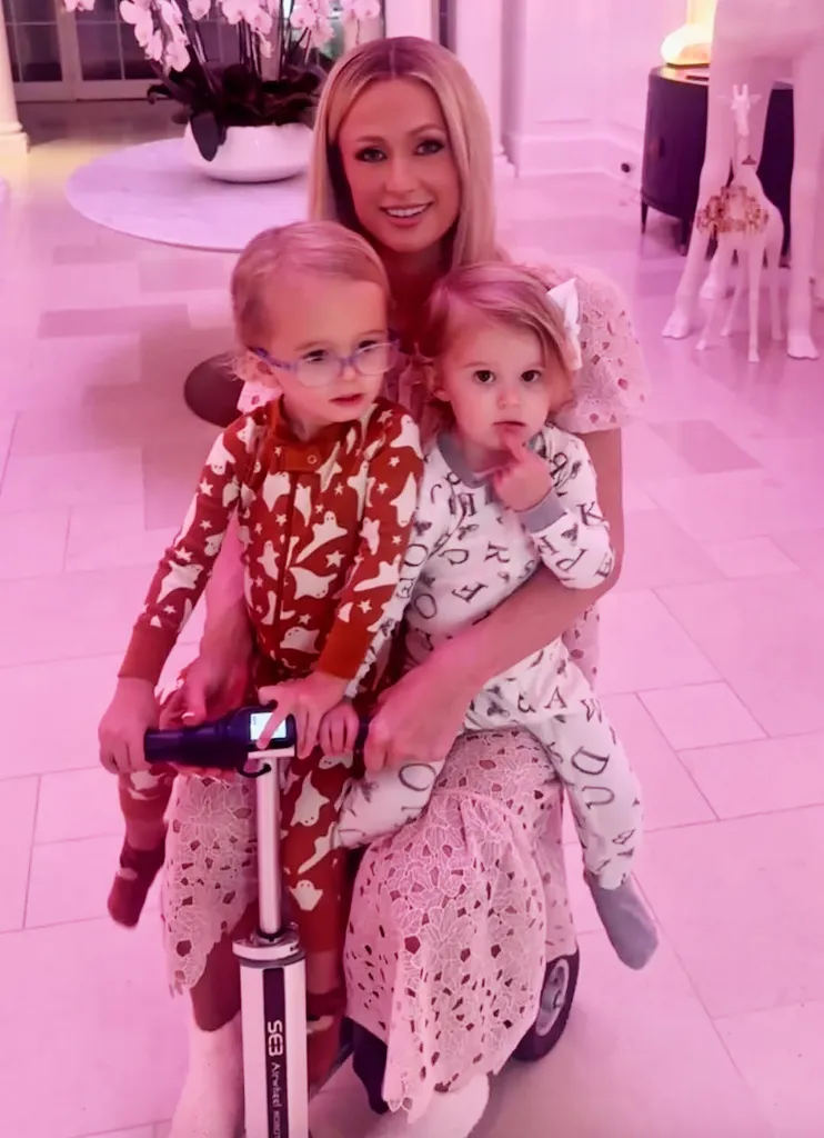 Paris Hilton smiling with her two children, a boy in a red ghost-patterned onesie on a scooter and a girl in a white letter-patterned onesie.