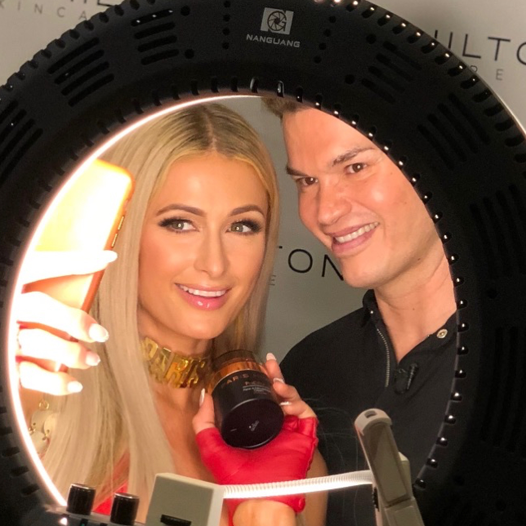 Paris Hilton and Kim Vo holding a skincare product and phone while looking into a ring light.