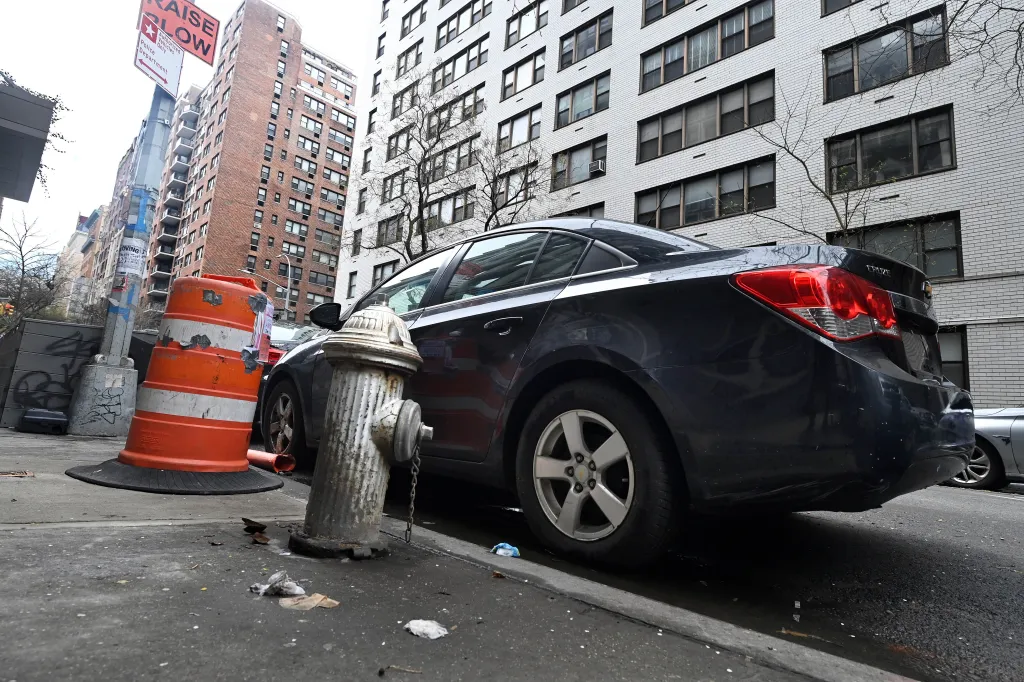 A dark blue car parked on a city street next to a fire hydrant and an orange construction barrel.