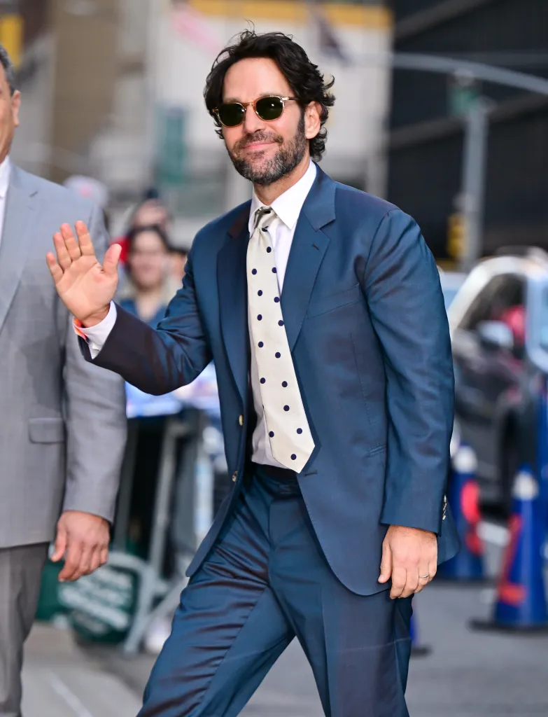 Paul Rudd in a blue suit, polka dot tie, and sunglasses waves to fans outside the Ed Sullivan Theater.