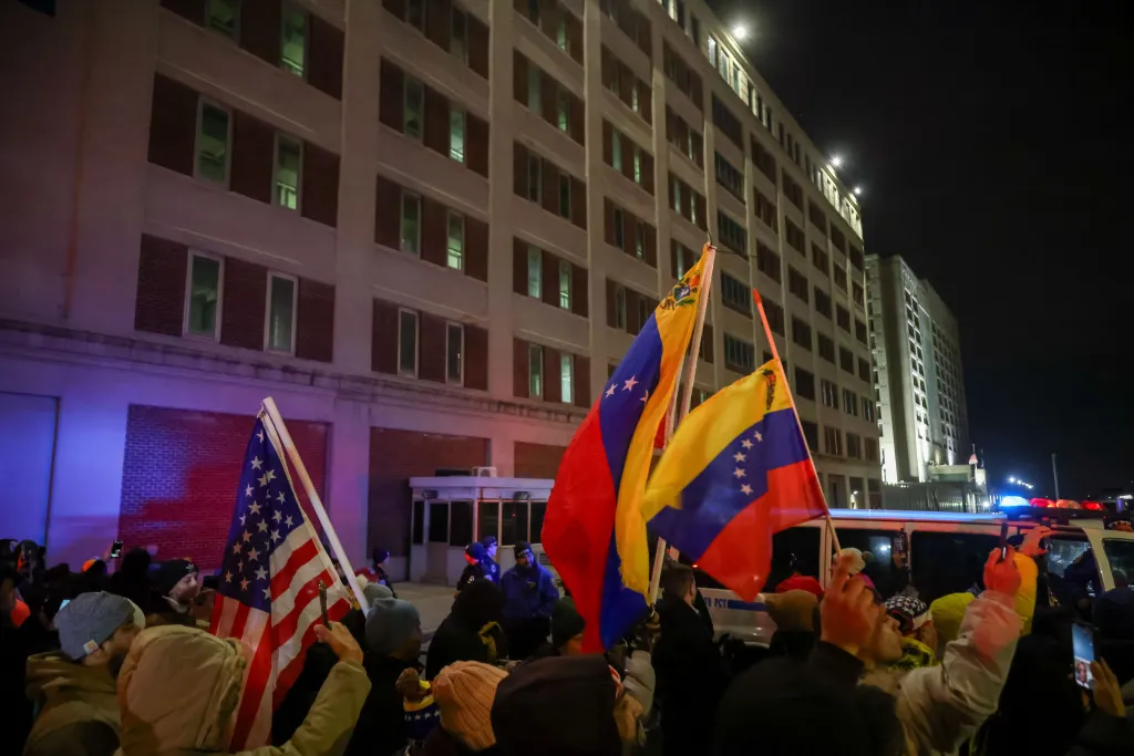 People holding Venezuelan and American flags celebrate outside of MDC Brooklyn at night.
