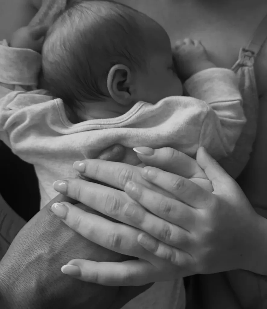 Black and white photo of a baby in a onesie held by two adults' hands, one with manicured nails.