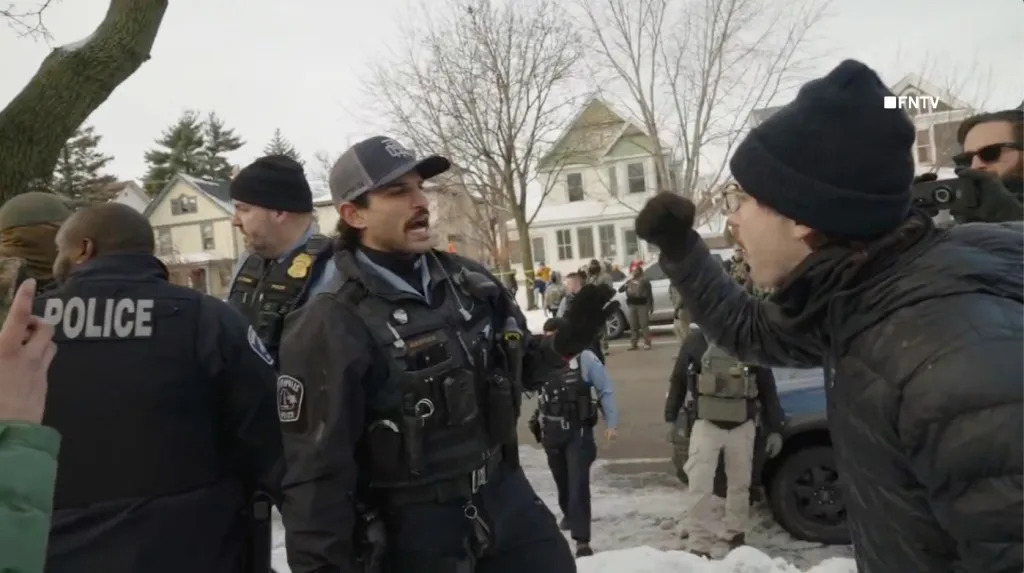 A police officer with a mustache and an activist in a black beanie yelling at each other in a snowy street.