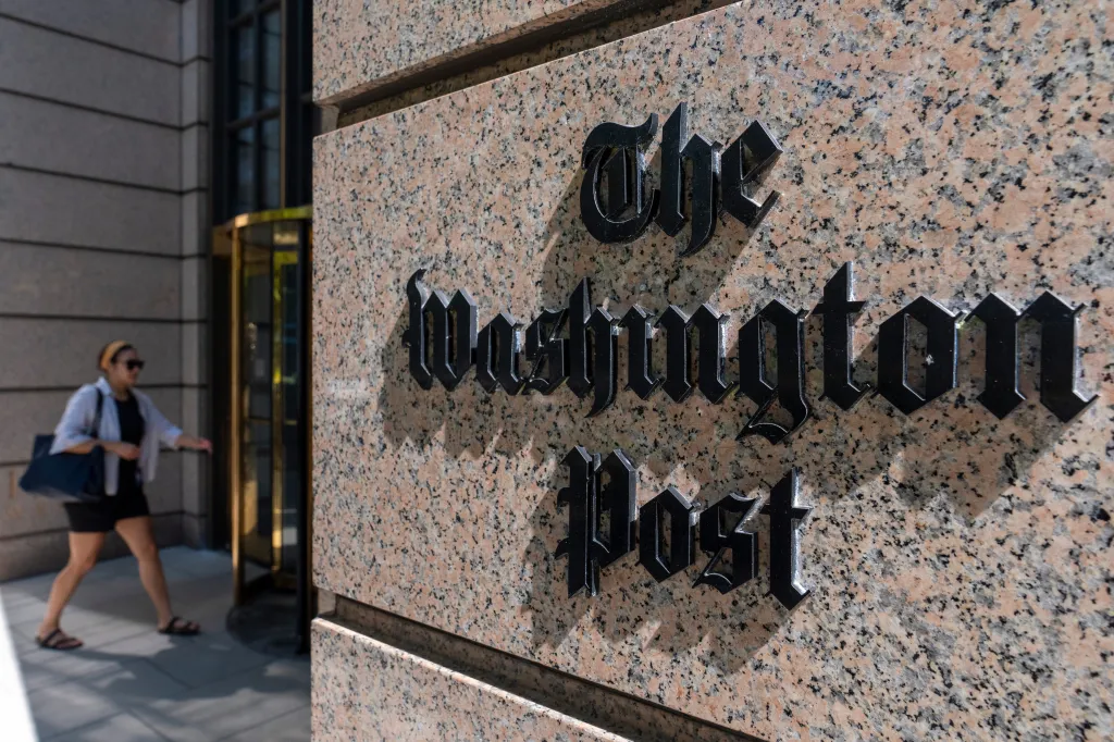 The Washington Post logo on the side of a building, with a person walking into the entrance in the background.