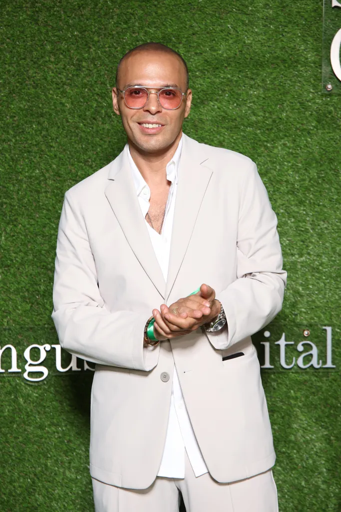 Richie Akiva in a light-colored suit, white shirt, and rose-tinted glasses.