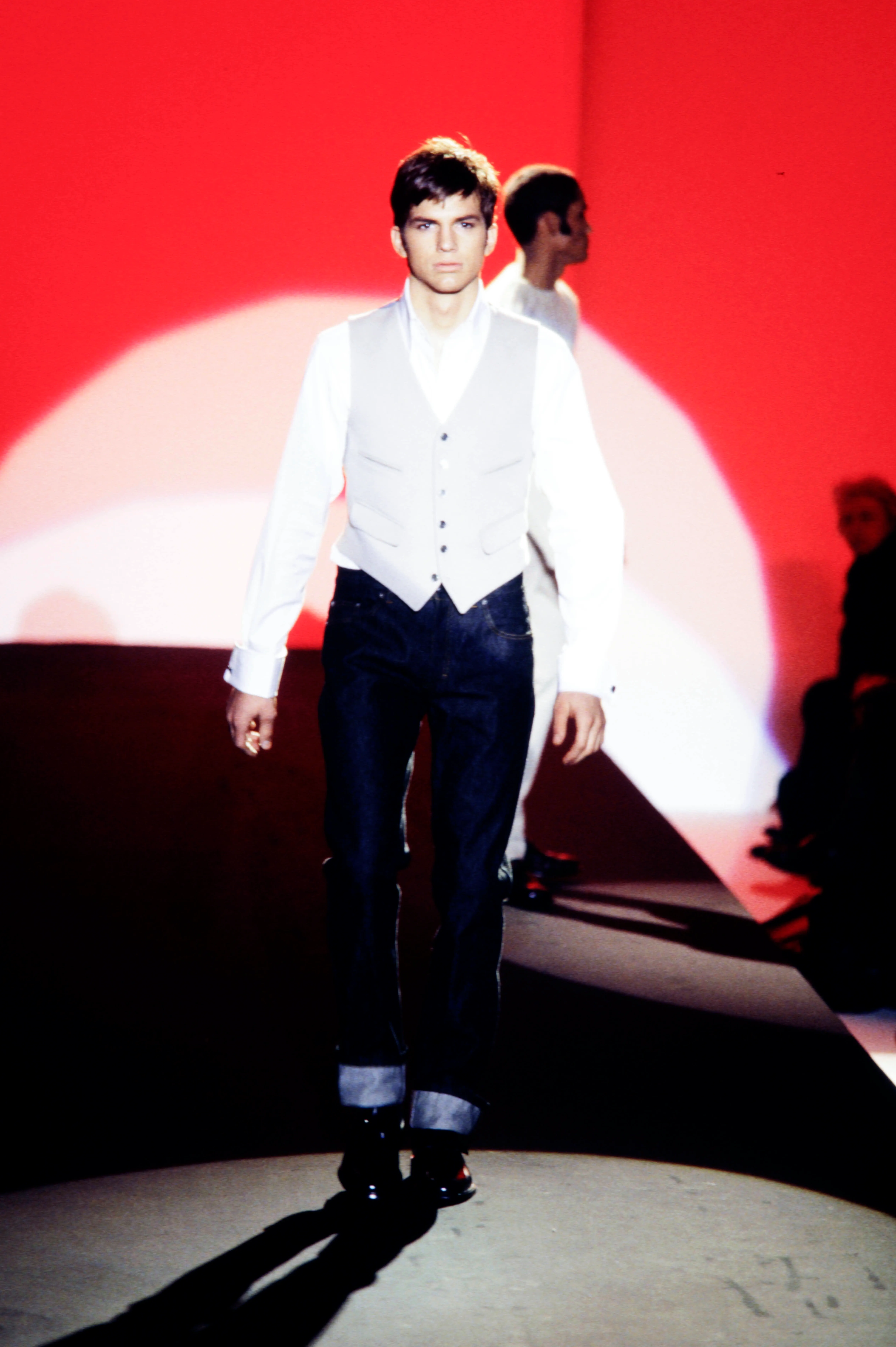 Ashton Kutcher walking the runway in a white shirt, light gray vest, and dark jeans with rolled cuffs.