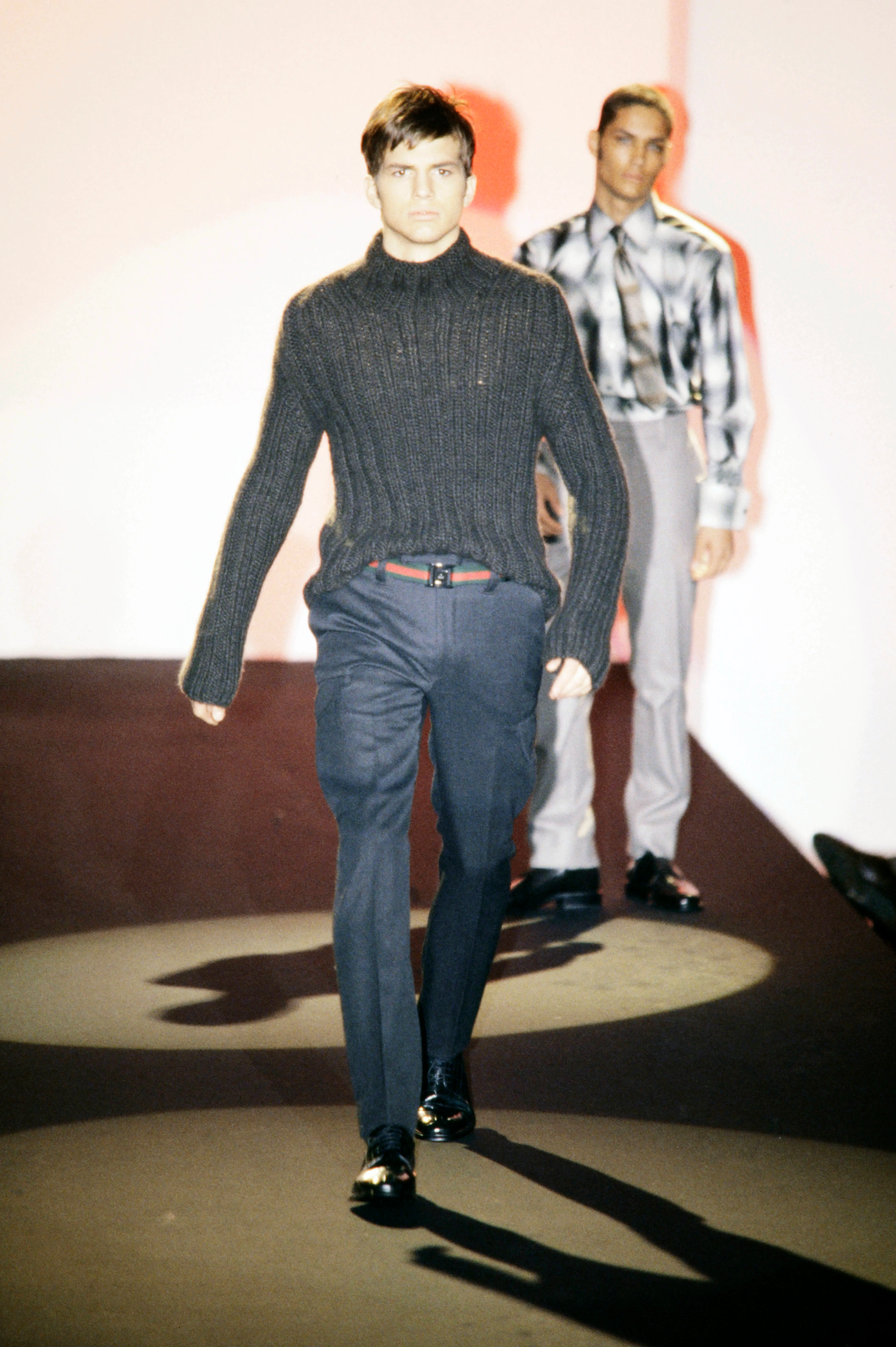 Ashton Kutcher modeling a dark grey knit turtleneck and dark trousers with a Gucci belt on the runway.