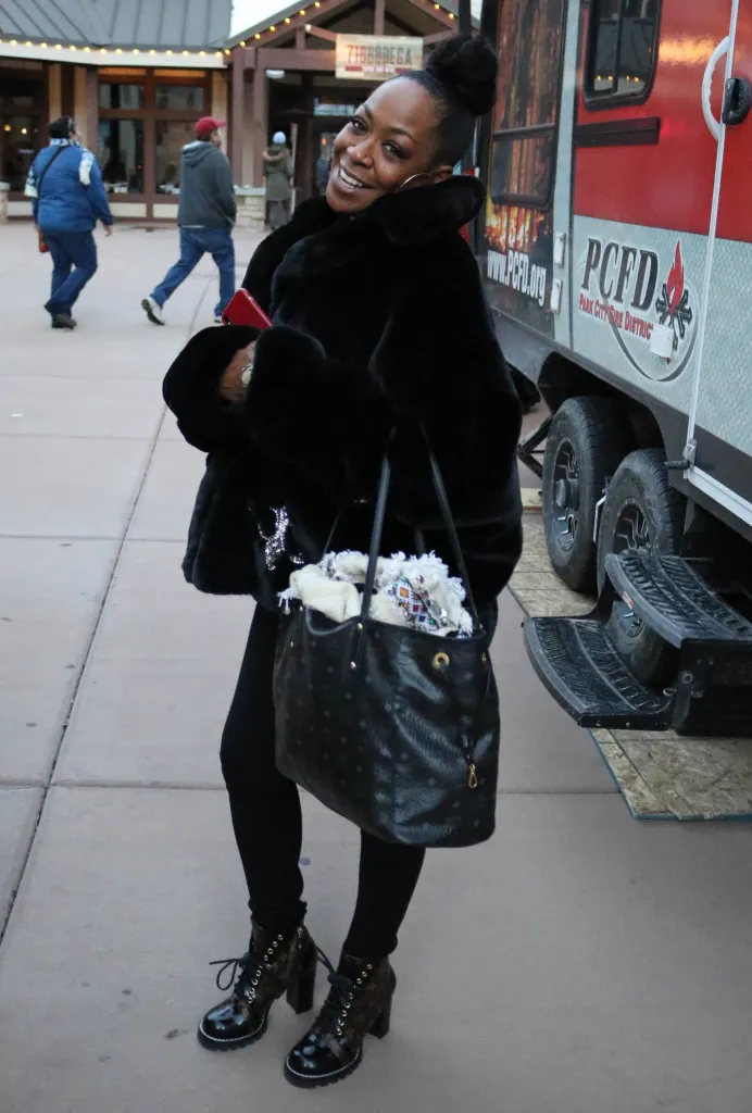 Tichina Arnold smiling at the camera, wearing a black fur coat and holding a black purse, next to a red trailer labeled 