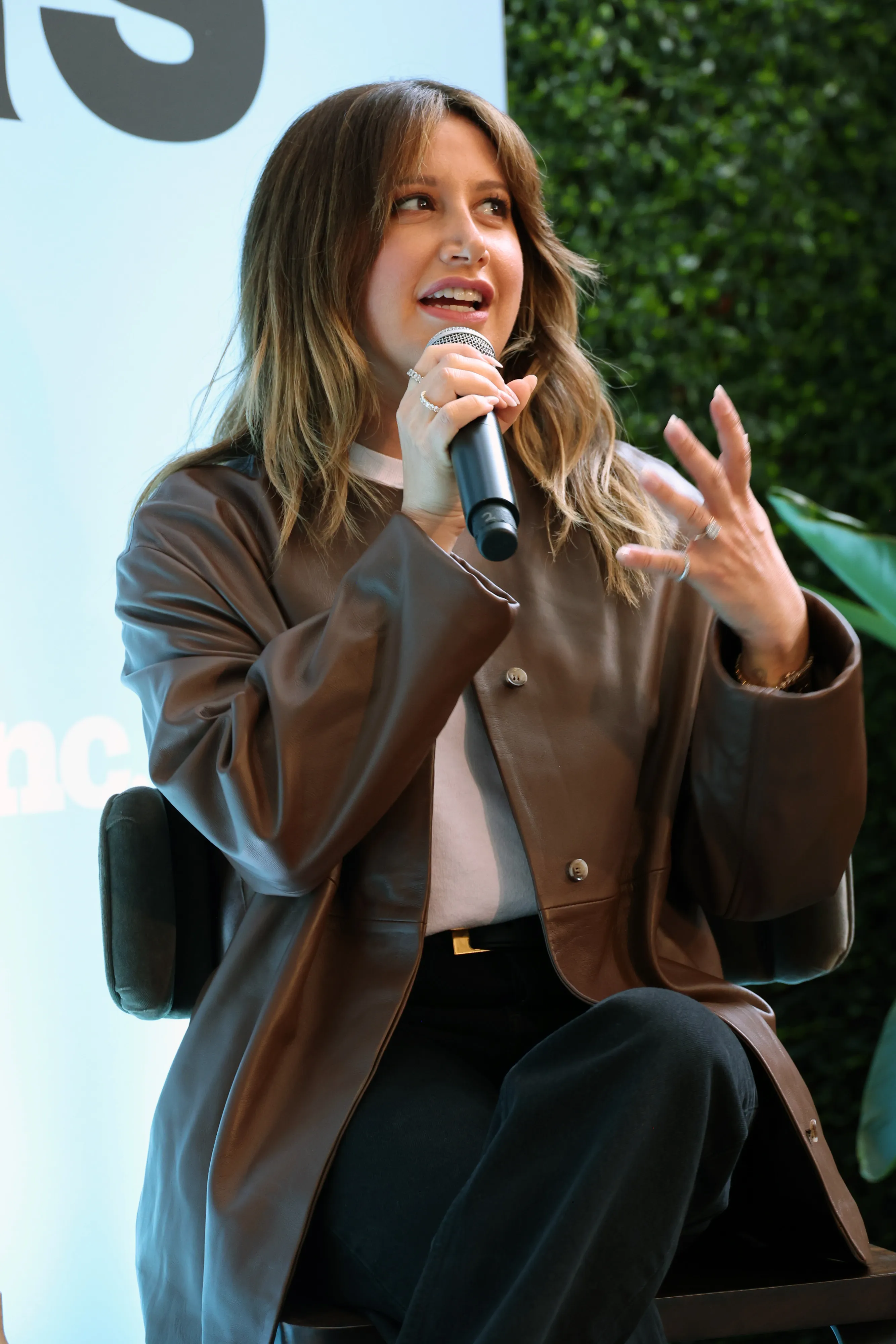 Ashley Tisdale speaking into a microphone while sitting in a chair, wearing a brown leather jacket.