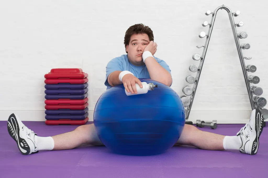Overweight man sitting on the floor with an exercise ball looking tired.