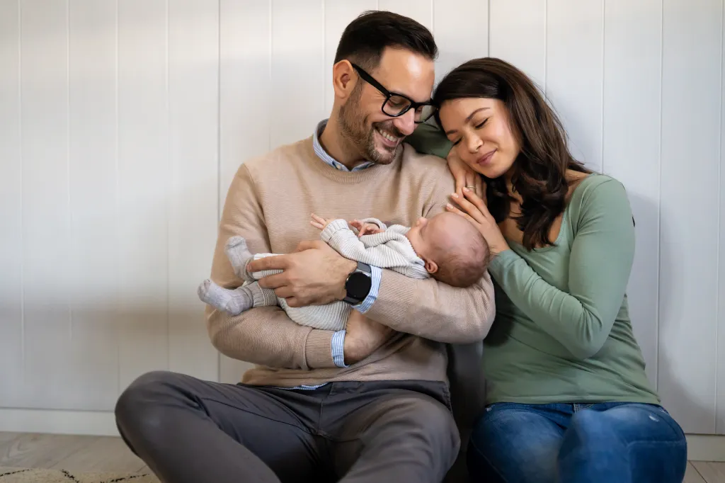 A happy couple sitting on the floor, the father holding their newborn baby, and the mother resting her head on the father's shoulder.