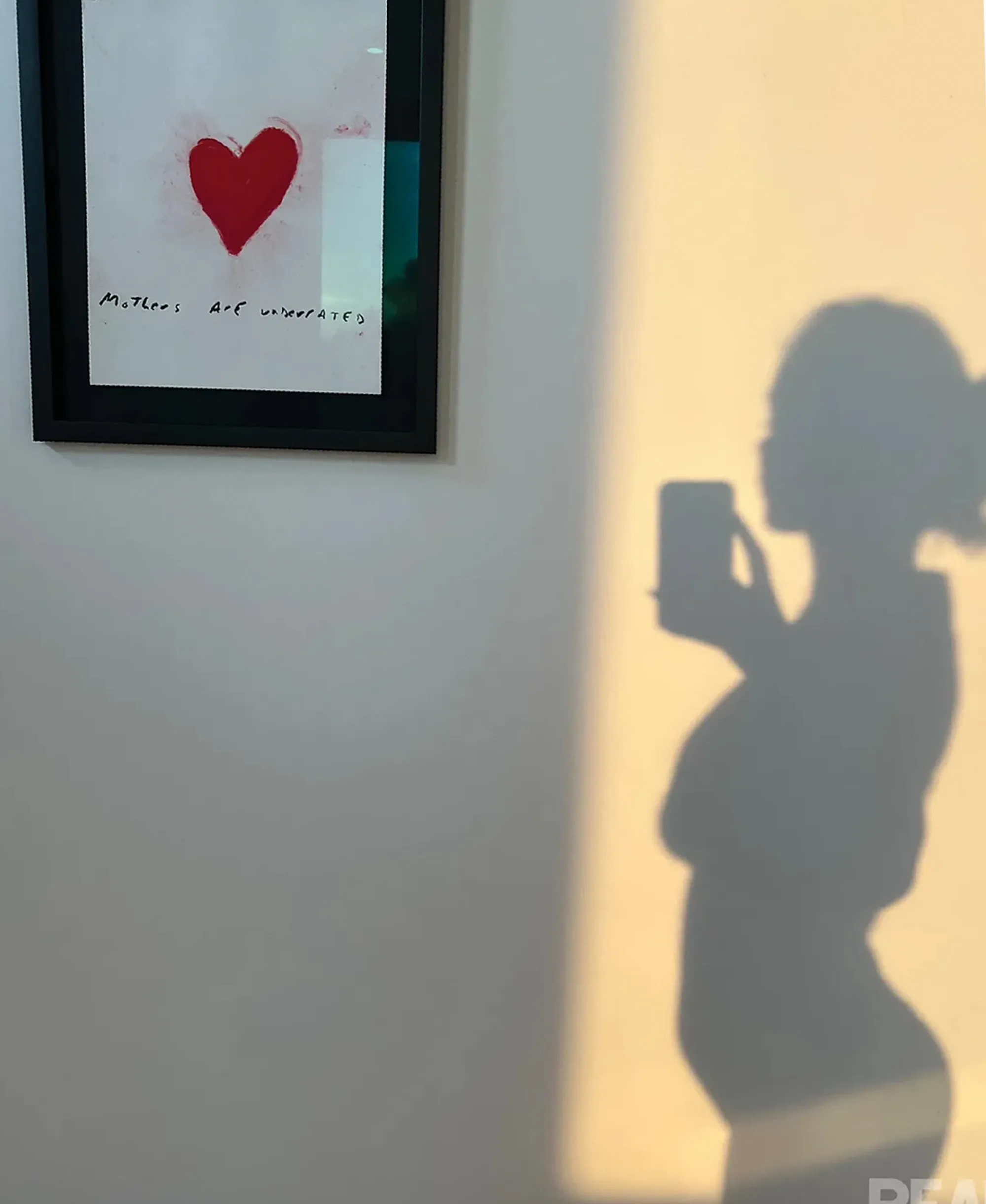 Shadow of a person with a baby bump taking a photo next to a framed picture of a red heart and the words 