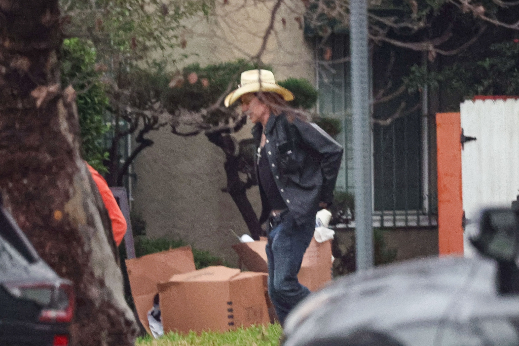 Mickey Rourke in a straw hat and black jacket walking past cardboard boxes on a grassy area.