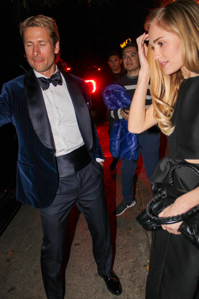 Glen Powell in a navy tuxedo jacket and Michelle Randolph in a black dress, with two men behind them.
