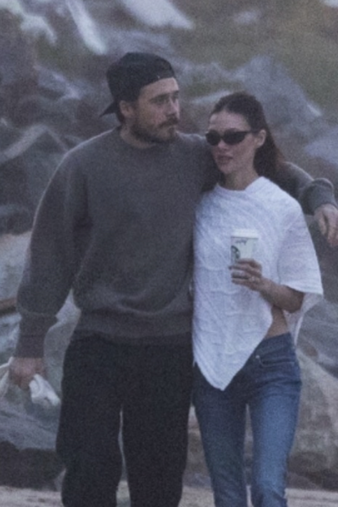 Brooklyn Beckham and Nicola Peltz Beckham walking arm-in-arm on a Malibu beach at sunset, Nicola holding a Starbucks cup.