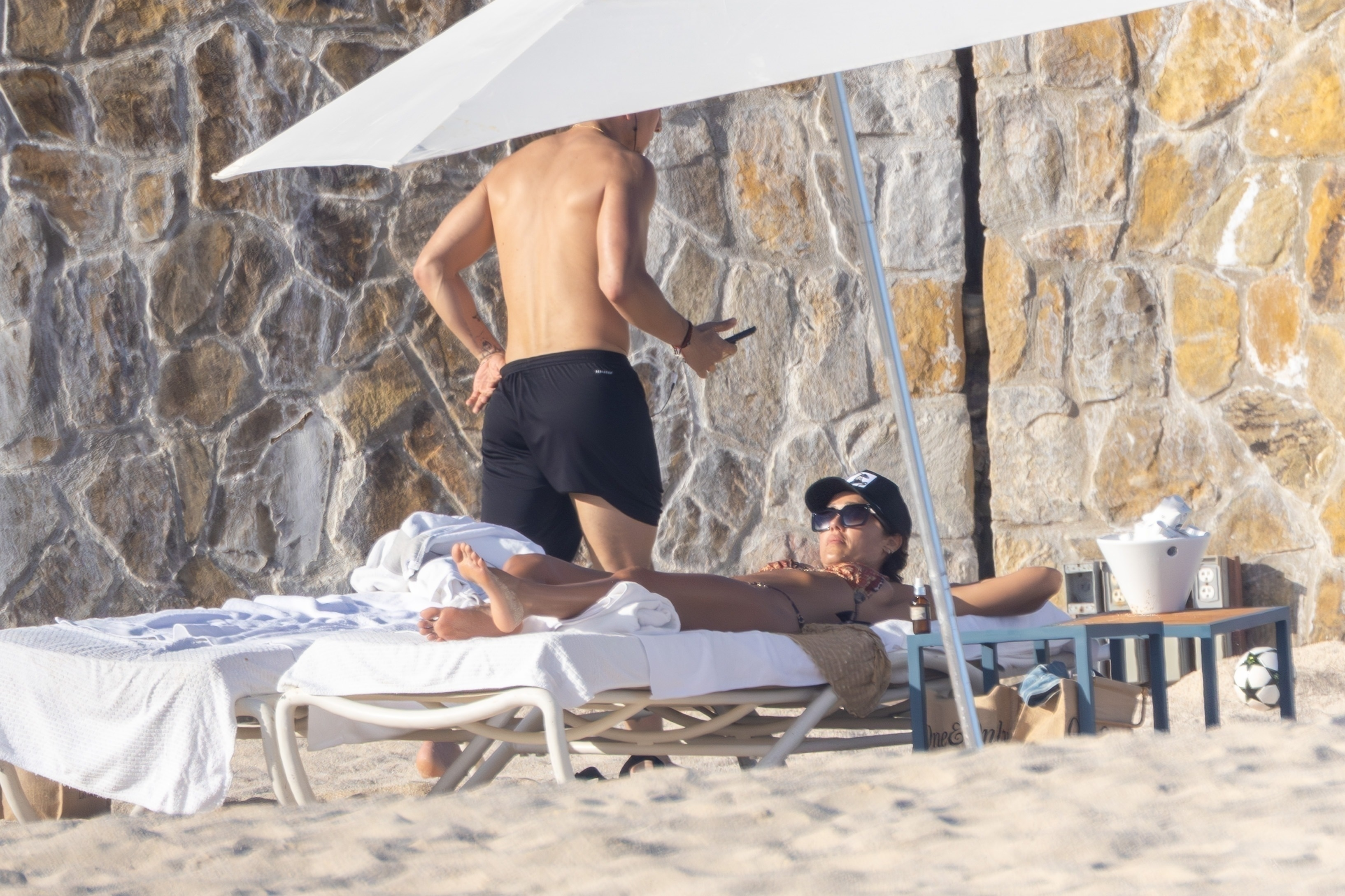 Jessica Alba relaxing on a beach lounge chair under an umbrella while Danny Ramirez stands beside her.