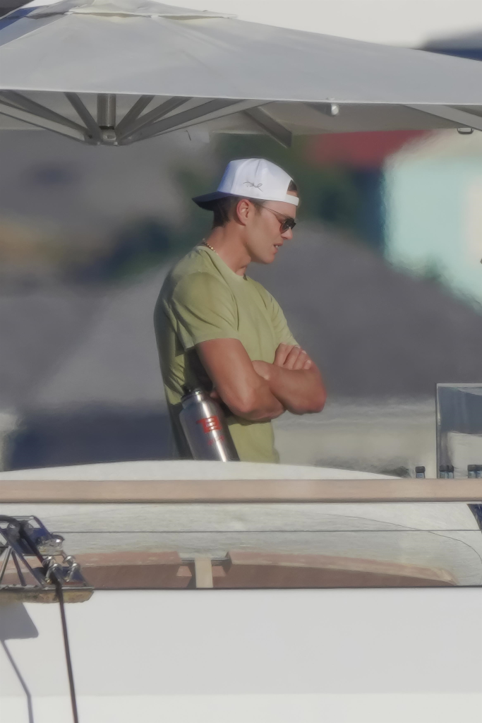 Tom Brady in a white baseball cap, sunglasses, and olive t-shirt, standing with his arms crossed on a yacht.