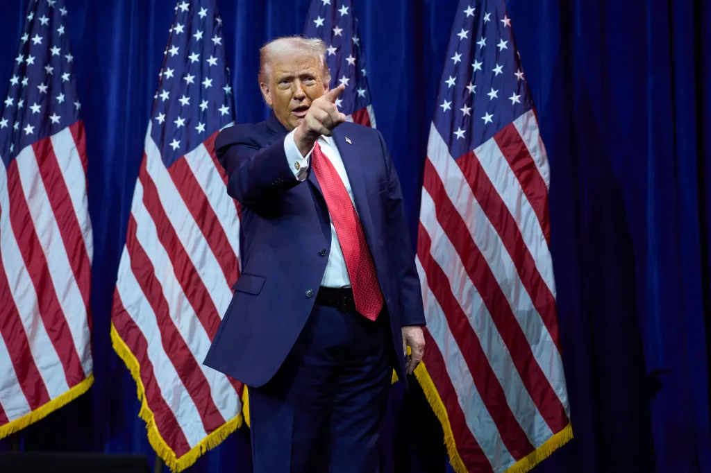President Donald Trump in a navy suit and red tie pointing while walking off stage, with American flags on either side.